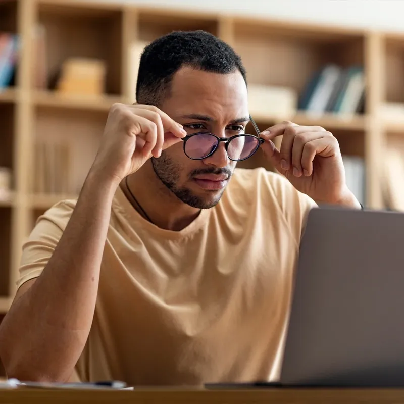 Man in a cardigan adjusting his glasses while reading at a laptop in warm light